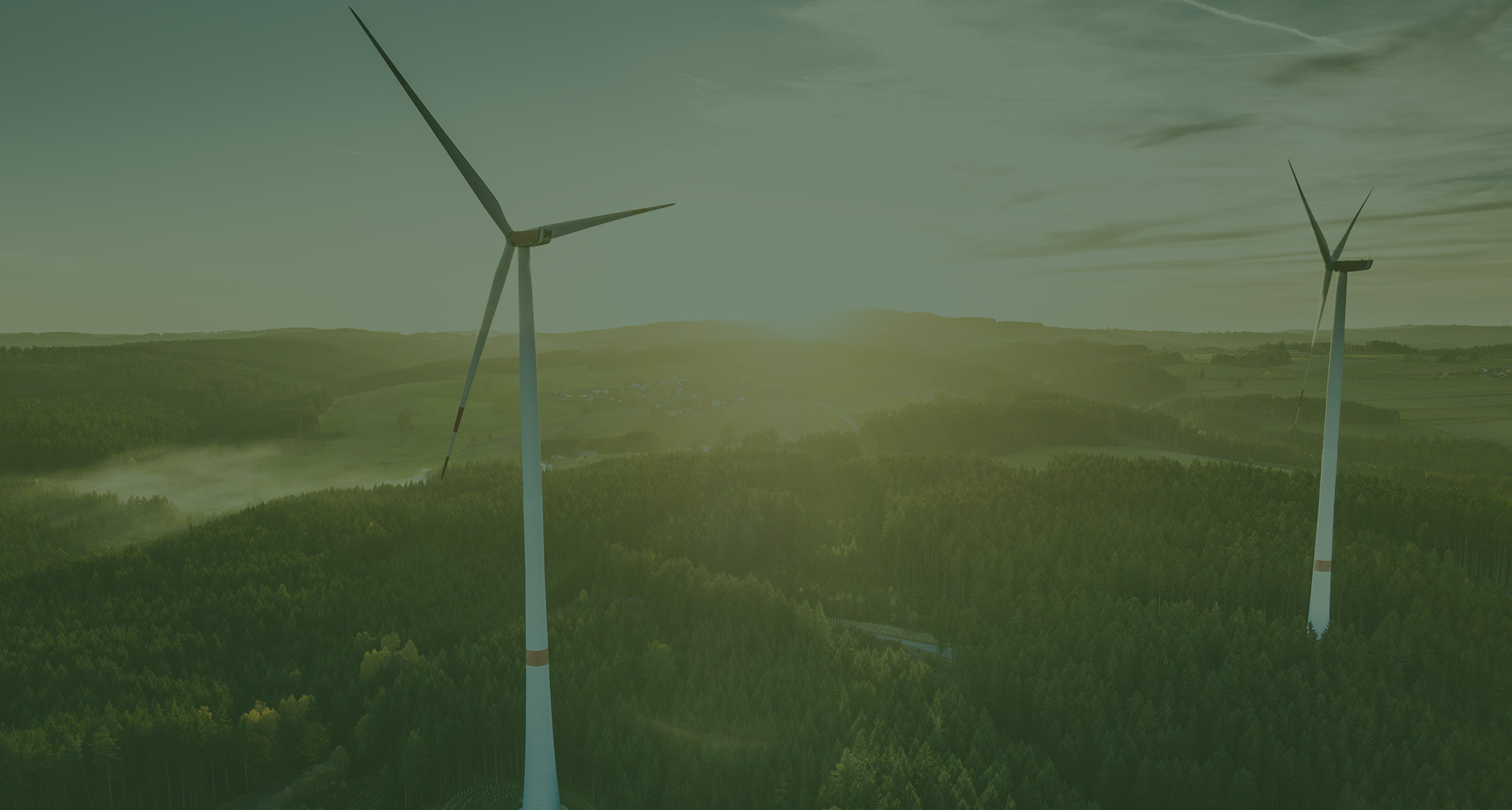 Rein dekoratives Element. Zu sehen ist ein Bild von zwei großen Windrädern, welche inmitten einer weitläufigen Waldfläche stehen. Man erkennt im Hintergrund die untergehende Sonne. Das Bild hat insgesamt ein grünes Overlay aus gestalterischen Zwecken.